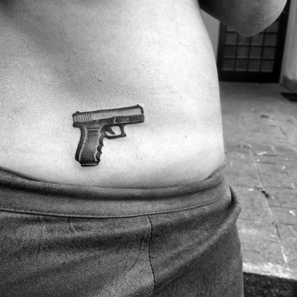 Small black and grey tattoo of a Glock pistol on the lower abdomen, featuring fine details and shading for a minimalist yet bold design.