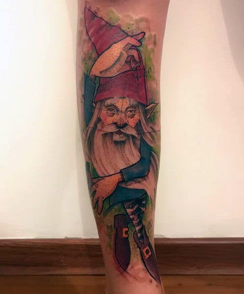 Tattoo of a whimsical gnome with a tall red hat, long white beard, and pointed shoes, surrounded by abstract green watercolor-style accents.
