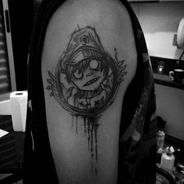 Sketch-style black ink tattoo of 2D from Gorillaz on the upper arm, with a crazed expression, dripping ink, and circular emblem framing him.