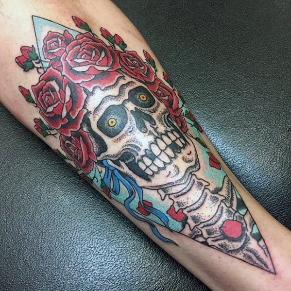 Grateful Dead skeleton tattoo with wide yellow eyes, framed by vivid red roses and a blue triangle background on the forearm.
