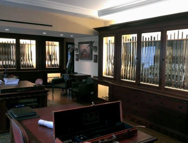 Elegant gun room with illuminated wood display cabinets, leather seating, and a classic desk for a refined, masculine style.