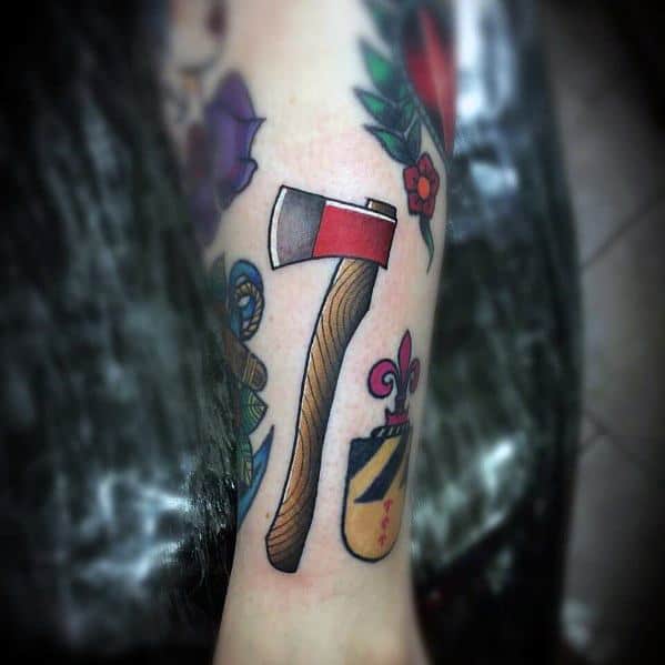 Traditional-style hatchet tattoo with a red and silver blade, a curved wood-textured handle, and bold outlines, surrounded by other colorful designs.