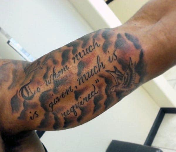 Script tattoo on the inner bicep reading 'To whom much is given, much is required,' with a swallow and flame-like shading in the background