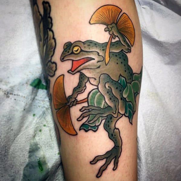 Japanese-style tattoo featuring a dynamic green frog holding two golden fans, perched on a branch with flowing leaves, symbolizing agility and tradition.