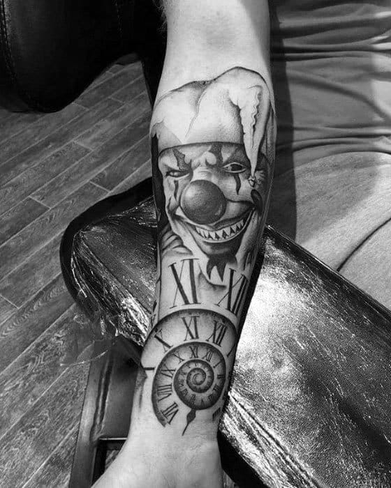 A black and gray tattoo of a sinister clown with a jester hat, surrounded by Roman numerals, a spiral design, and a clock, placed on the forearm.