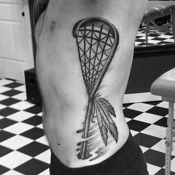 Black and grey lacrosse stick tattoo with intricate netting, feathers, and flowing lines, placed on the ribs for a bold and symbolic design.