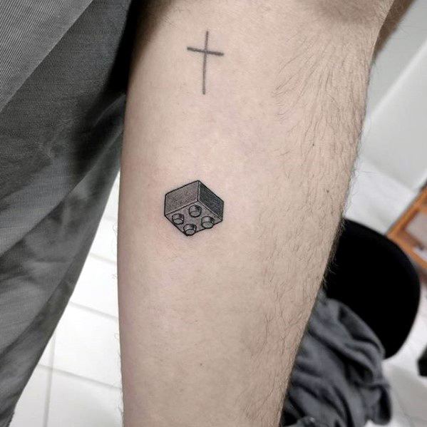 Small black and grey tattoo of an isometric Lego brick with subtle shading, placed below a simple cross on the forearm.