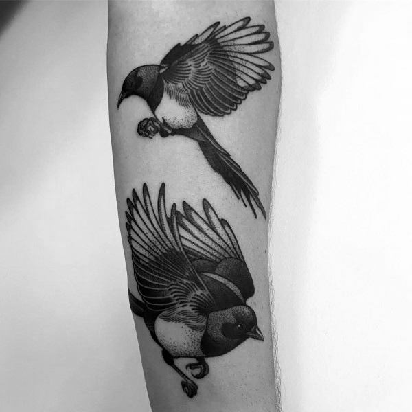 Black and gray magpie tattoo on forearm, featuring two birds in flight with intricate shading and detailed feathers for a dynamic composition.