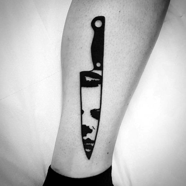 Minimalist black ink tattoo of a knife with Michael Myers’ face subtly reflected in the blade, creating a bold horror design.