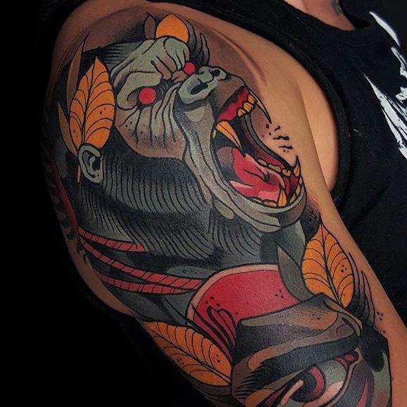 Neo-traditional gorilla tattoo on upper arm with red eyes, open mouth, tribal elements, and bold orange and yellow leaves.