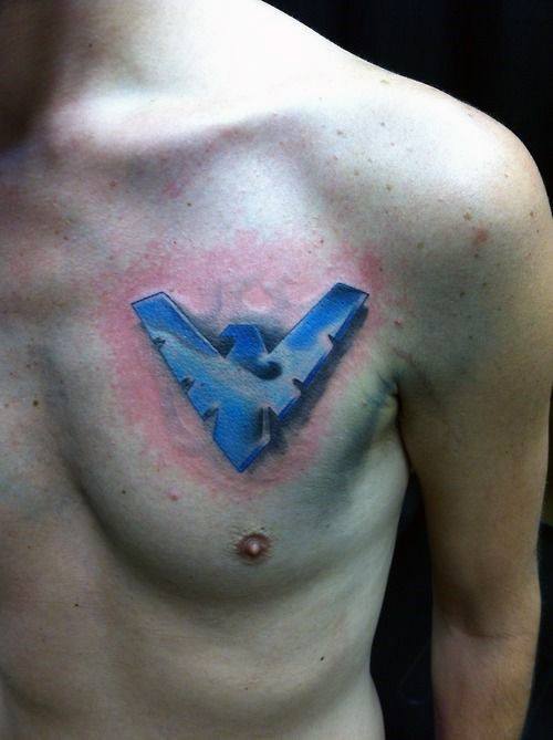 Nightwing emblem tattoo on the chest with a 3D effect in blue and black tones, giving the illusion it's embedded in the skin.