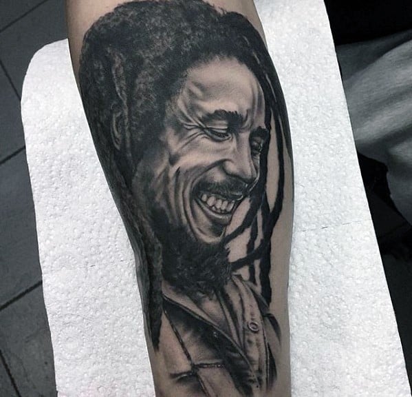 Forearm tattoo of Bob Marley smiling with detailed dreadlocks and textured shading, done in a realistic black and gray style.