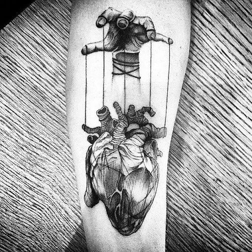 Black and grey tattoo of an anatomical heart controlled like a puppet by a hand, symbolizing manipulation and emotional vulnerability.