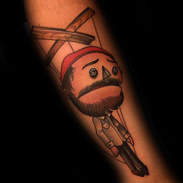 Cartoon-style puppet tattoo of a bearded man with a red beanie controlled by strings, giving a playful yet melancholic vibe.