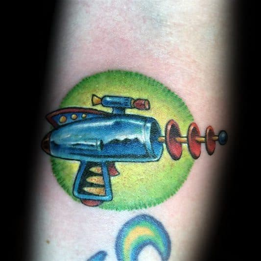Colorful retro ray gun tattoo with a metallic blue finish, set against a vibrant green background, featuring classic sci-fi-inspired details.