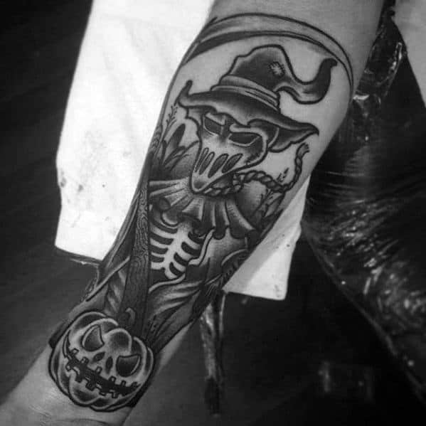 Bold blackwork scarecrow tattoo with a skeletal chest, sinister mask, witch hat, chain, and grinning jack-o'-lantern at the bottom.