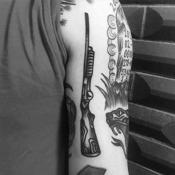 Black and grey shotgun tattoo on the upper arm, featuring bold outlines and wood grain detailing for a classic firearm design.
