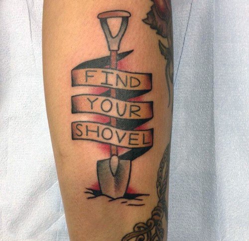 Shovel tattoo with a ribbon wrapped around it displaying the phrase "Find Your Shovel," symbolizing determination and perseverance.