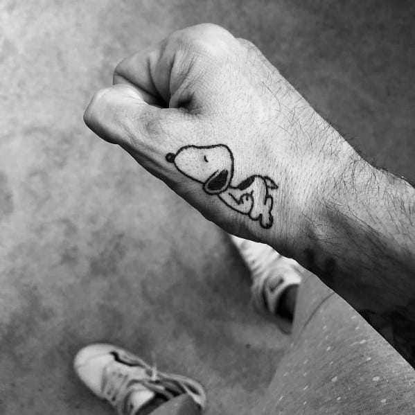 Black and white tattoo of Snoopy in a relaxed pose, placed on the back of a person's hand.