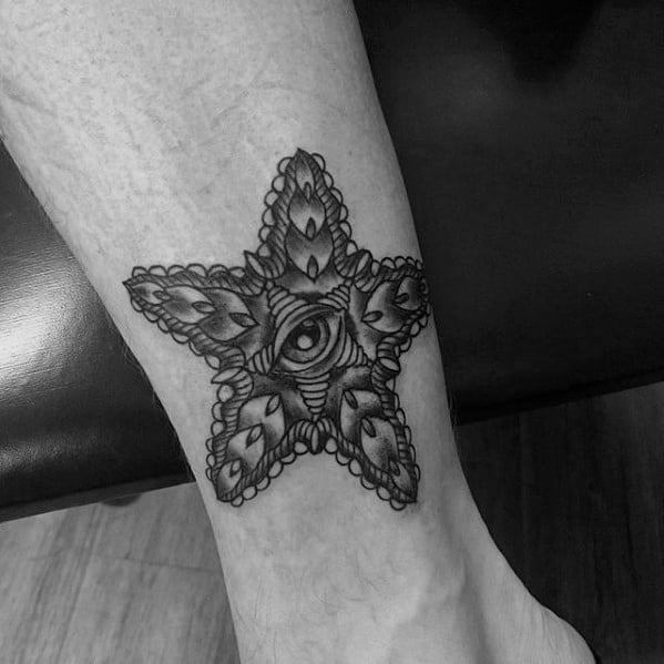 Black ink starfish tattoo with ornamental patterns and an all-seeing eye in the center, placed on the inner ankle.