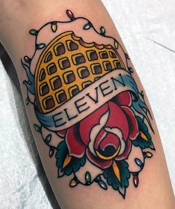 Traditional-style tattoo of a bitten waffle with a banner reading “Eleven,” surrounded by a rose and Christmas lights.