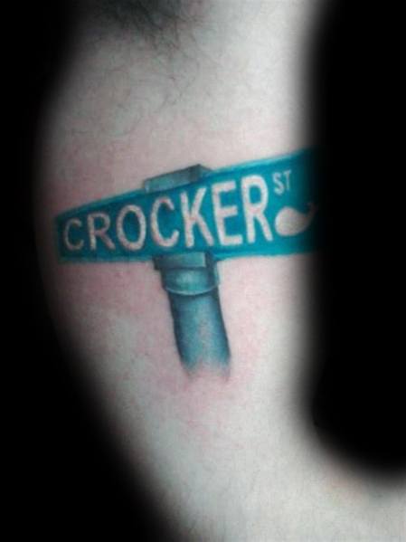 Color tattoo of a blue 'Crocker St' street sign with realistic shading and a subtle whale icon, placed on the upper arm.
