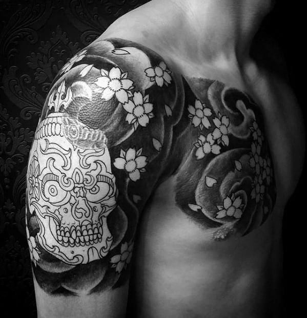 Black and grey Tibetan skull tattoo on shoulder and chest with floral accents and swirling background for a bold, contrasting look.