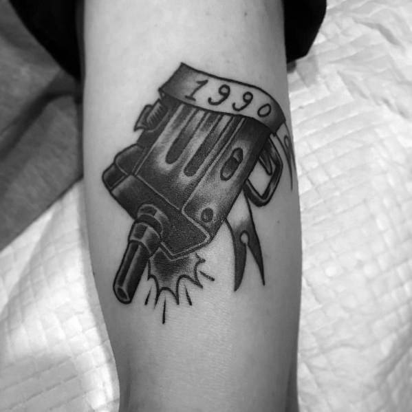 Black and grey Uzi tattoo with a ribbon wrapped around it displaying "1990," featuring bold shading and a stylized gunfire effect.