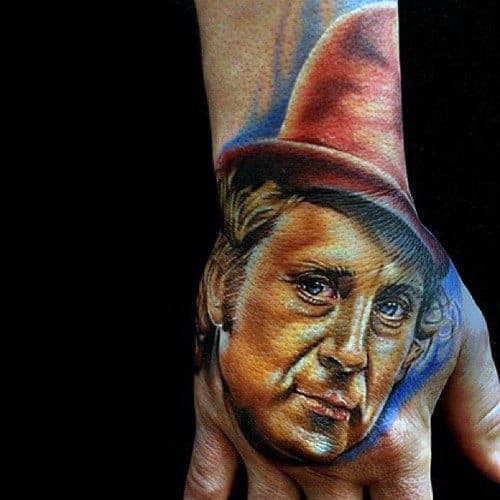 Realistic Willy Wonka tattoo on the hand, featuring warm tones, a red top hat, and a thoughtful expression with vivid shading.
