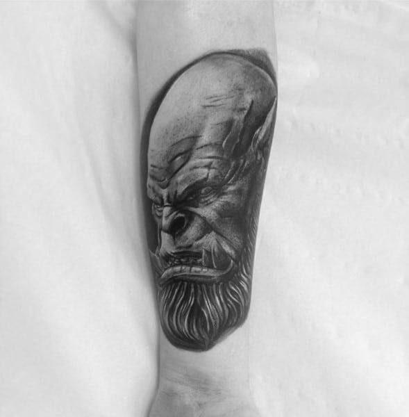 Realistic black and grey tattoo of an orc from World of Warcraft, featuring sharp details, fierce expression, and a long beard, inked on forearm.