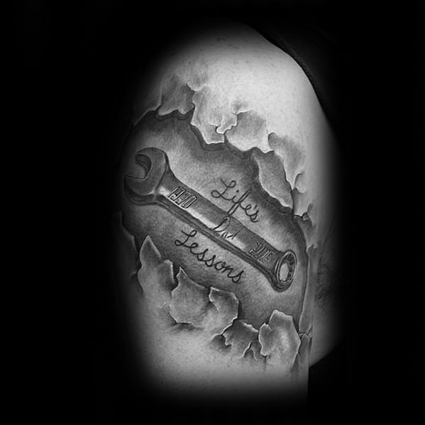Black and grey tattoo of a wrench beneath torn skin effect, with the words Life’s Lessons written across the tool in cursive script.