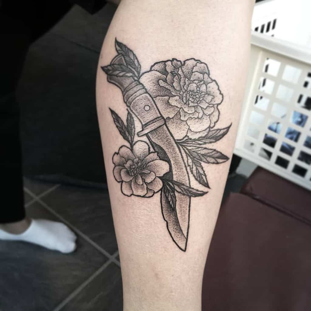 Black and grey tattoo featuring a detailed dagger entwined with blooming marigolds and leaves, with fine dot work and shading