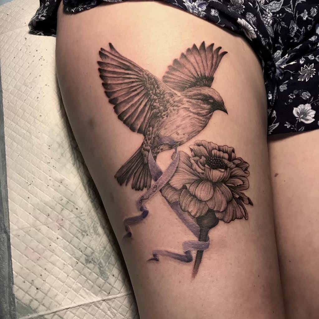 Realistic black and grey tattoo featuring a detailed bird in flight holding a flowing ribbon with "R. O", paired with a beautifully shaded marigold flower