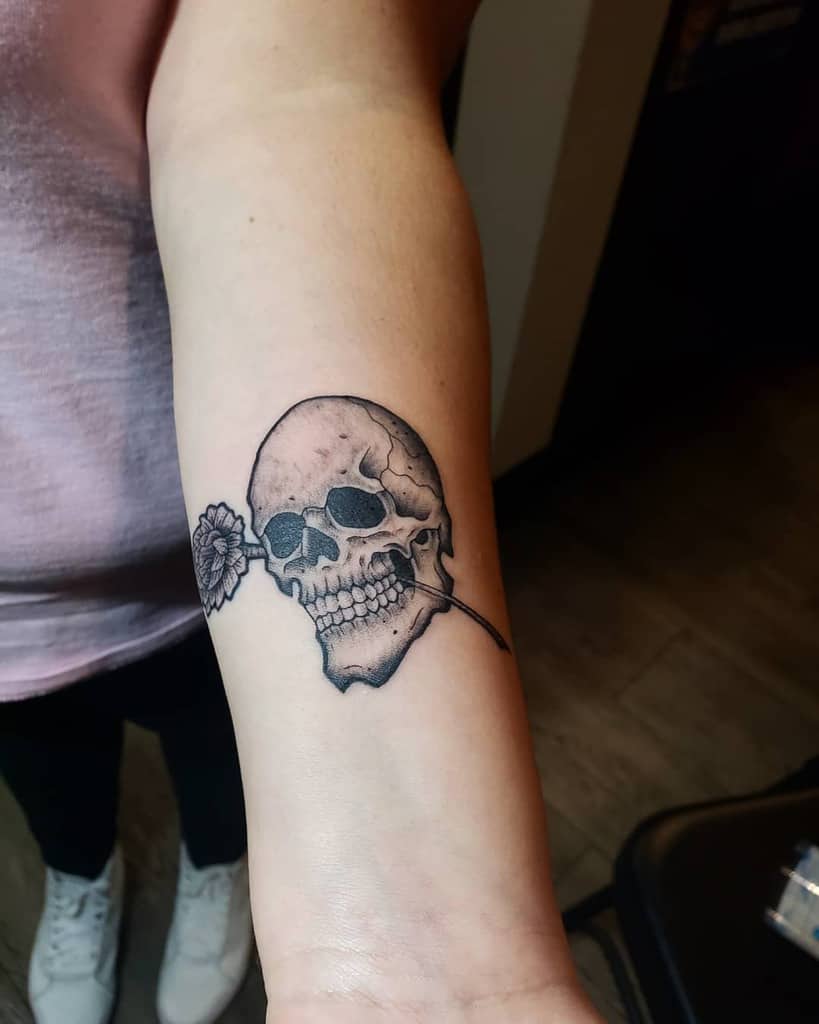 Black and grey tattoo of a detailed skull holding a marigold flower in its teeth, featuring fine shading and bold line work