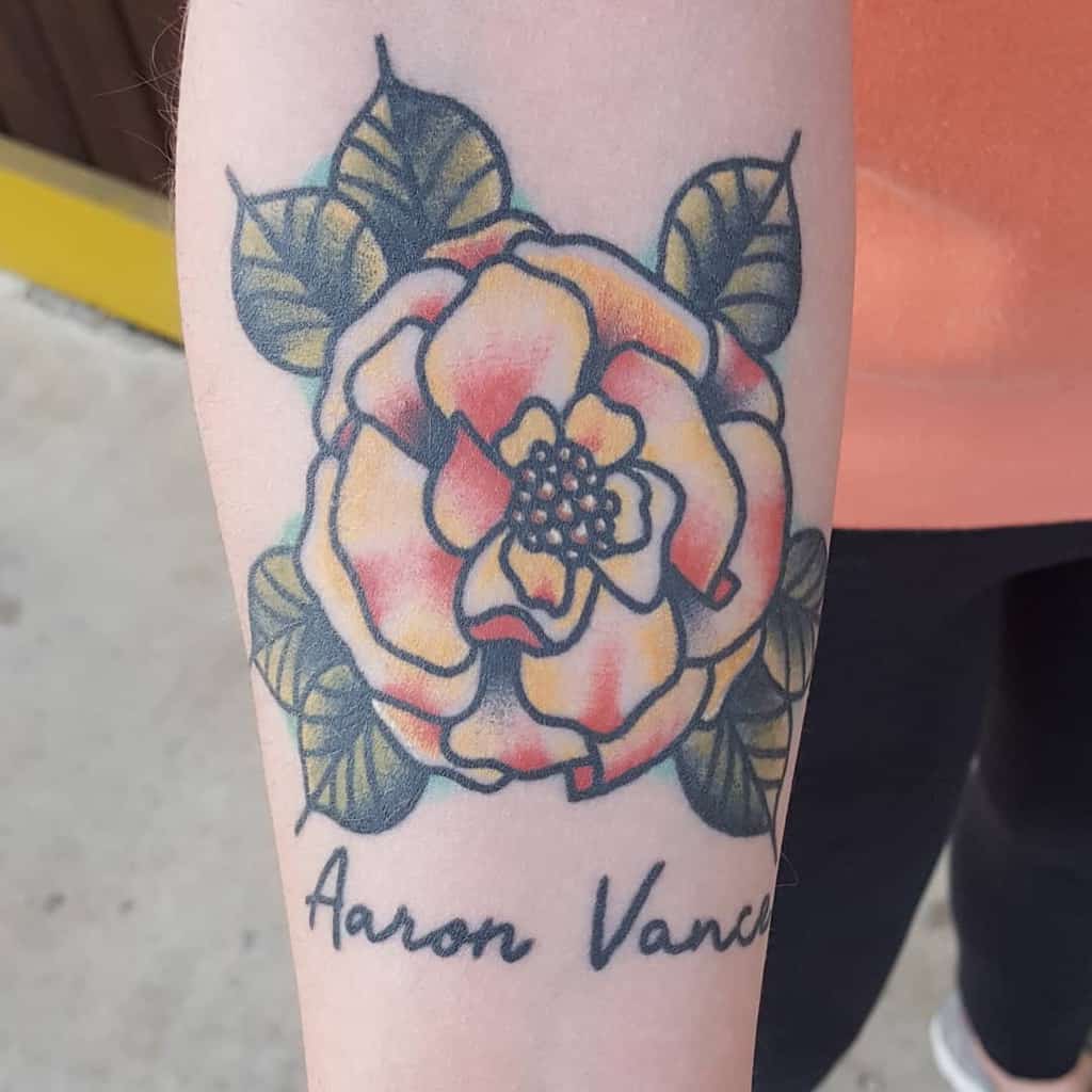 Tattoo featuring a stylized orange marigold with bold outlines, green leaves, and the name 'Aaron Vance' scripted below
