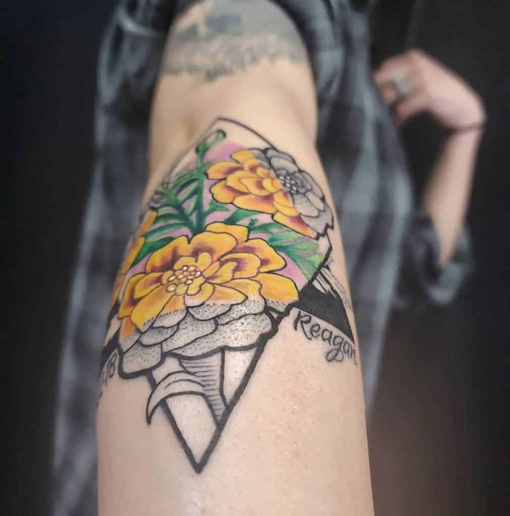 Colorful tattoo featuring vibrant orange marigolds growing from a geometric cracked design, with the name 'Reagan' scripted below.