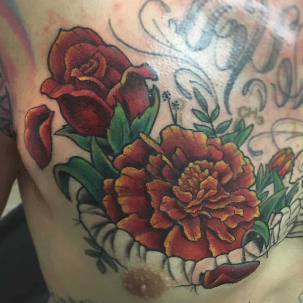 Bold tattoo featuring a deep red rose and a vibrant orange marigold blooming from a cracked surface, with rich shading and green leaves