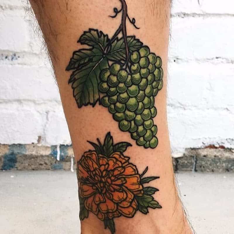 Tattoo of green grapes with vine and leaves above an orange marigold with leaves