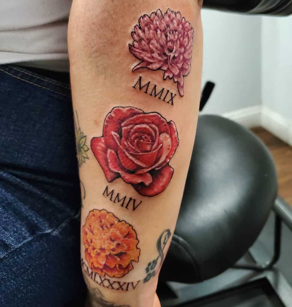 Tattoo featuring three detailed flowers, a chrysanthemum, rose, and marigold, each paired with Roman numerals representing significant years