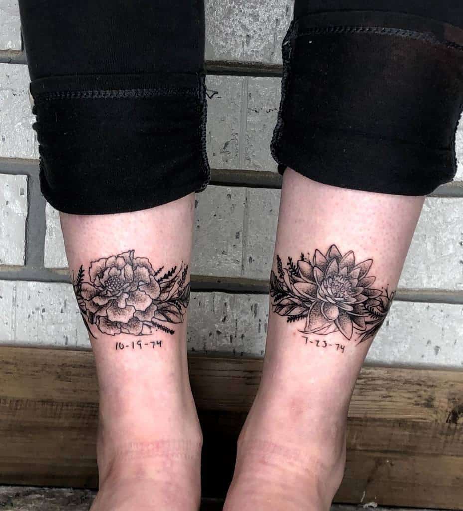 Matching black and grey tattoos featuring a marigold on one leg and a lotus on the other, framed by leaves and dates '10-19-74' and '7-23-14'