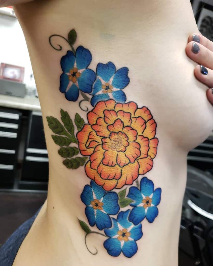 Colorful tattoo featuring a bright orange marigold surrounded by vivid blue flowers and lush green leaves in a bold, vibrant design