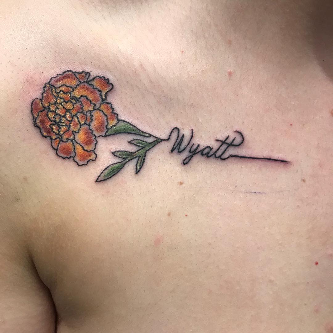 Minimalist tattoo featuring an orange marigold with green leaves, seamlessly blending into the cursive name 'Wyatt' as part of the stem