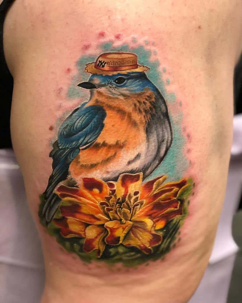 Colorful tattoo featuring a bluebird wearing a straw hat perched on a vibrant orange marigold, with a soft watercolor-style background