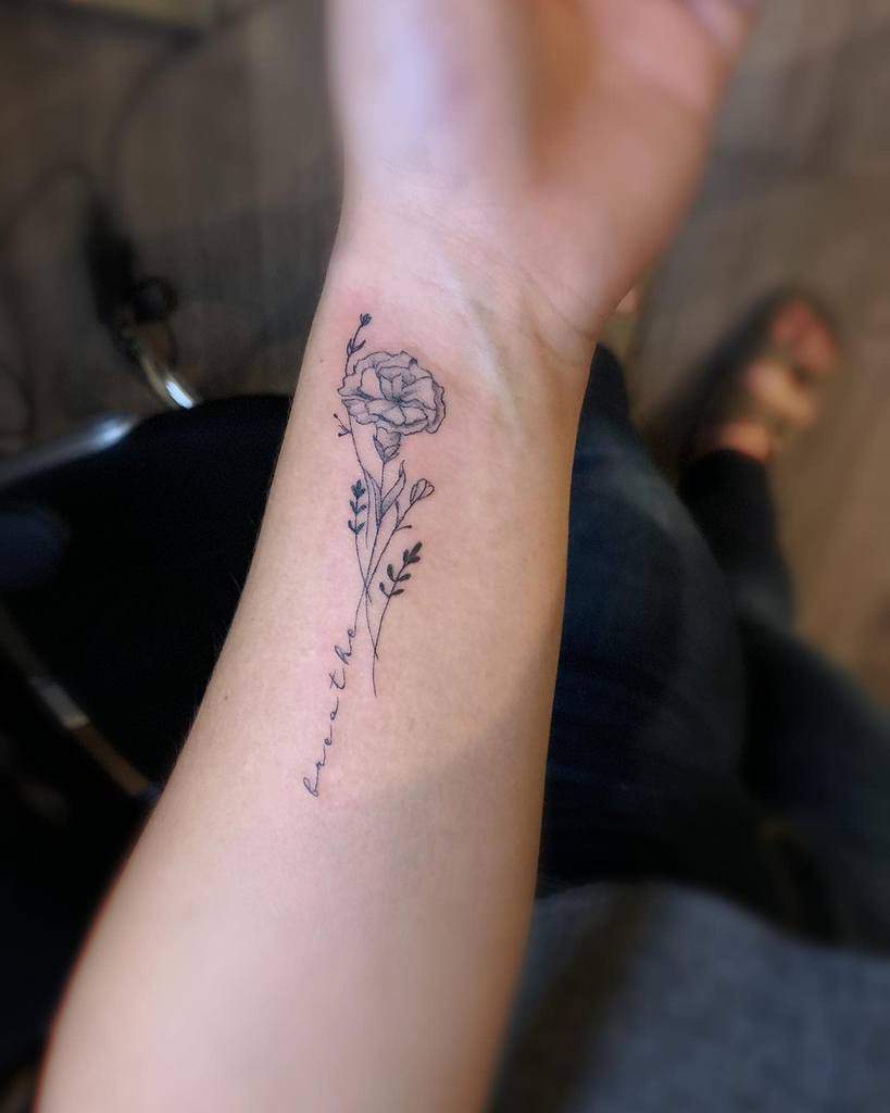 A tattoo of a delicate marigold with thin stem and leaves, accompanied by cursive text "Breathe" on the side