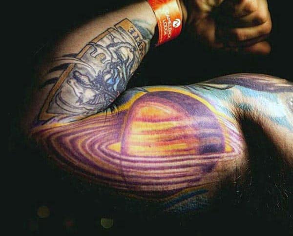 Vibrant tattoo of Saturn with glowing rings in shades of purple, orange, and yellow, creating a cosmic effect on the inner bicep