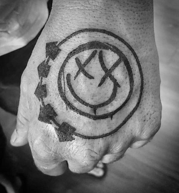 Black and white tattoo of the Blink-182 smiley face logo with arrows, placed on the back of the hand with bold black lines.