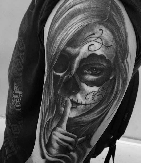 La Catrina tattoo of a woman with sugar skull makeup, one darkened eye socket, and a finger over her lips in a shushing gesture.