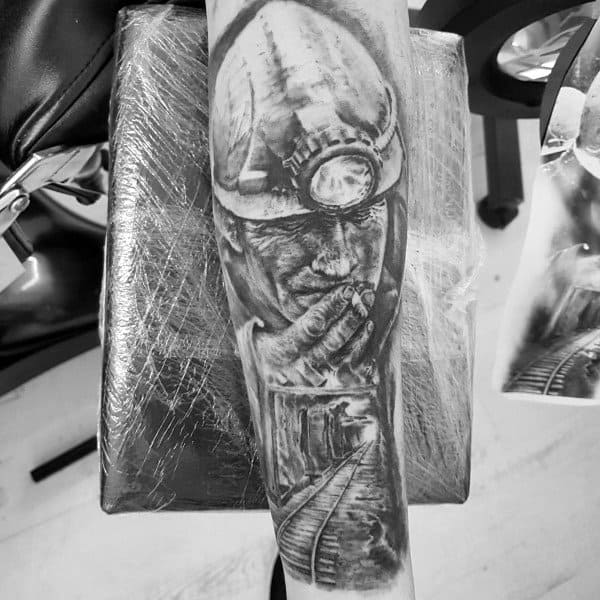 Black and grey tattoo of a reflective coal miner with a helmet and headlamp, holding his face above a dark mining tunnel.