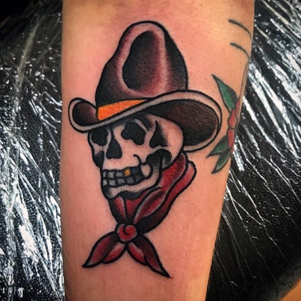 Tattoo of a grinning cowboy skull wearing a brown hat and red bandana, done in a bold traditional style with strong outlines and shading.