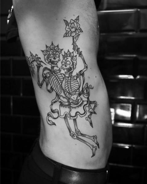 Elegant dancing skeleton tattoo featuring two skeletons in traditional attire, holding floral staffs, creating a mystical and artistic design on the ribs.